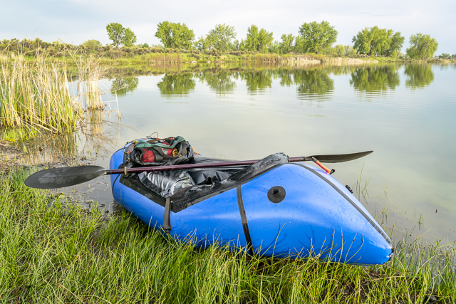 a blue pacraft (one-person light raft used for expedition or adventure racing) with a kayak paddle on a lake shore in spring scenery