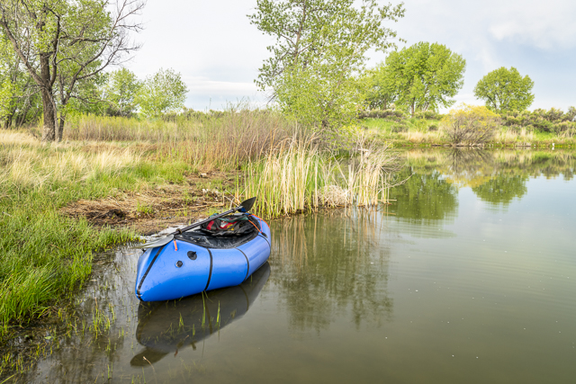 a blue packraft (one-person light raft used for expedition or adventure racing) with a kayak paddle on a lake shore in spring scenery