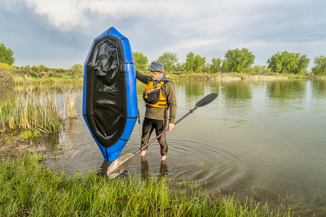a senior male with a blue pacraft (one-person light raft used for expedition or adventure racing) on a lake shore in spring scenery