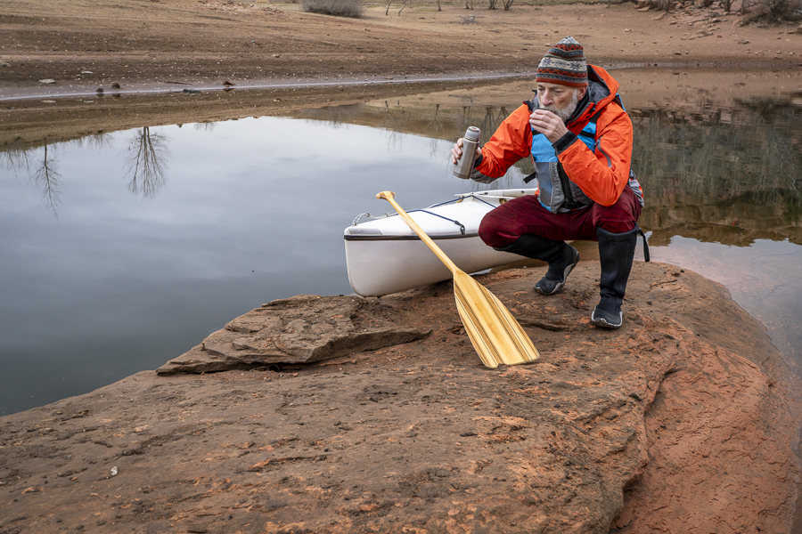 hot tea break - senior male paddler with a decked expedition canoe with a wooden paddle on a rocky shore of Horsetooth Reservoir near Fort Collins, Colorado, low water level fall or winter scenery