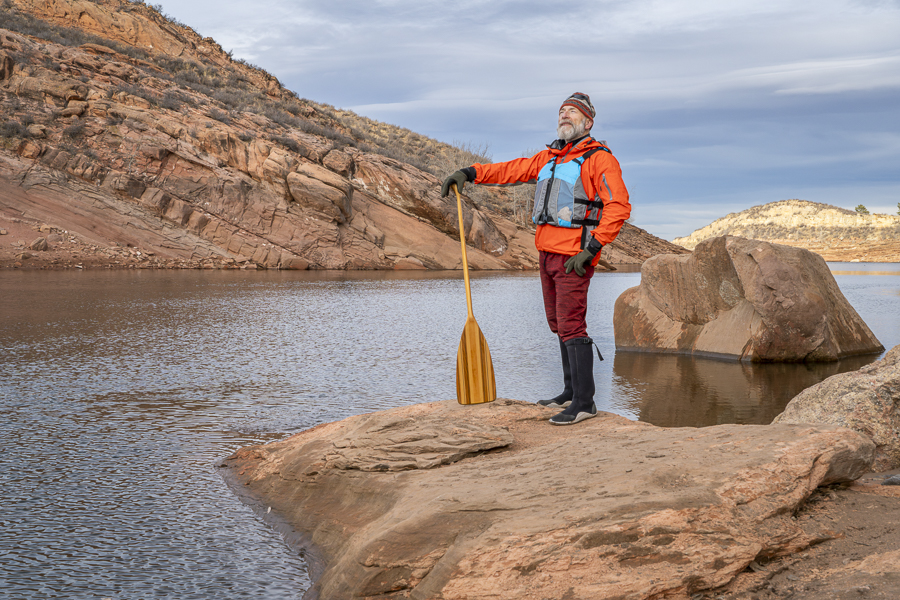 senior male paddler with a wooden canoe paddle on a rock shore of a mountain lake - Horsetooth Reservoir in fall or winter scenery