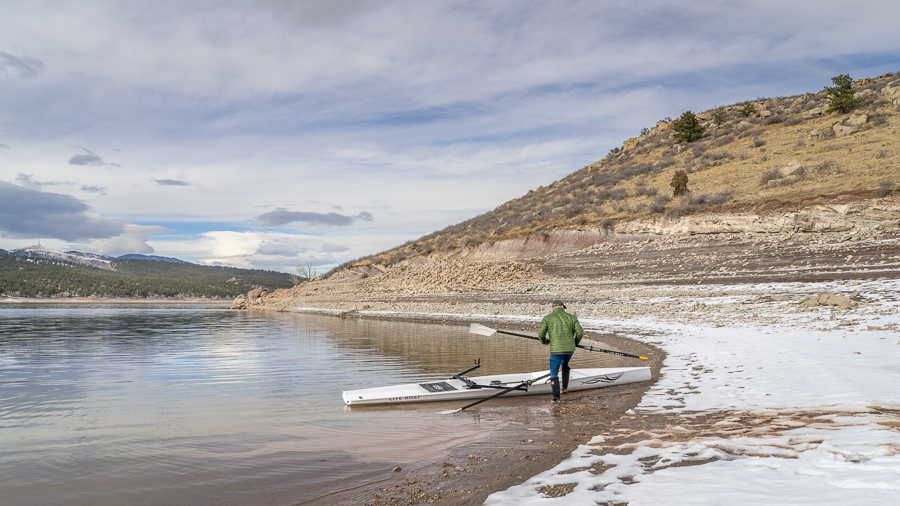 Senior male rower with a coastal rowing shell and hatchet oars on a shore of Carter Lake in winter scenery in northern Colorado.