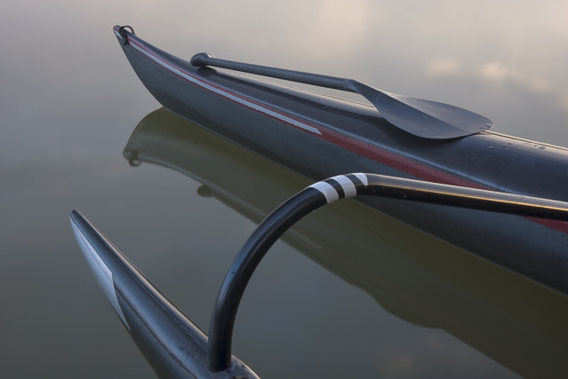 Carbon fiber bent shaft paddle on a slim bow of racing outrigger canoe, calm lake.