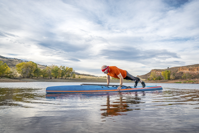 A senior male exercising (pushups and planks) on a stand up paddleboard, a calm mountain lake in fall colors