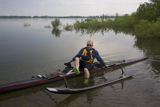 mature, male paddler in life jacket and green clogs is boarding his narrow black (carbon fiber) racing outrigger canoe, calm lake, springtime